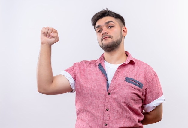 Jonge knappe kerel die roze poloshirt draagt die ok met vuist doet die ernstig status over witte muur kijkt