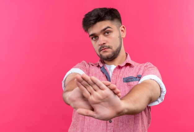 Jonge knappe kerel die roze poloshirt draagt die serieus stop met gekruiste handen doet die zich over roze muur bevinden