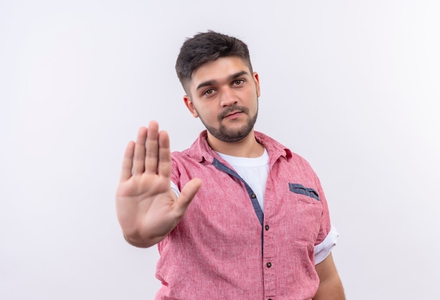 Jonge knappe kerel die roze poloshirt draagt die stopbord met hand doet die zich over witte muur bevindt