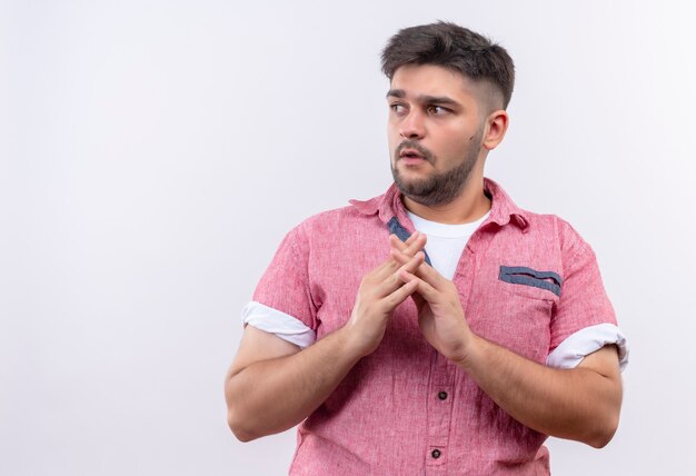 Jonge knappe kerel die roze poloshirt draagt en er bovendien gevaarlijk uitziet met samengevoegde handen die zich over een witte muur bevinden