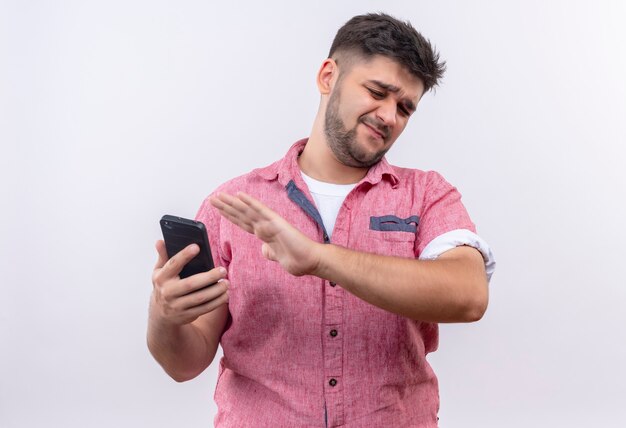 Jonge knappe kerel die roze poloshirt draagt en weigert te kijken naar de telefoon die zich over een witte muur bevindt