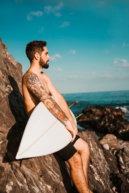 Jonge knappe kerel die surfplank dichtbij stenen houdt