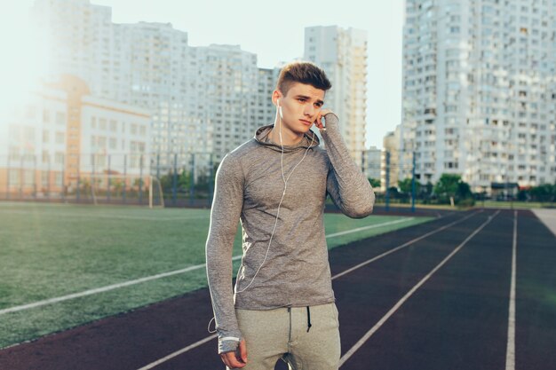 Jonge knappe kerel in grijs sportkostuum op atletiekbaan op gebouwenachtergrond in de ochtend. Hij draagt een grijs sportpak, een koptelefoon. Hij kijkt naar de kant.