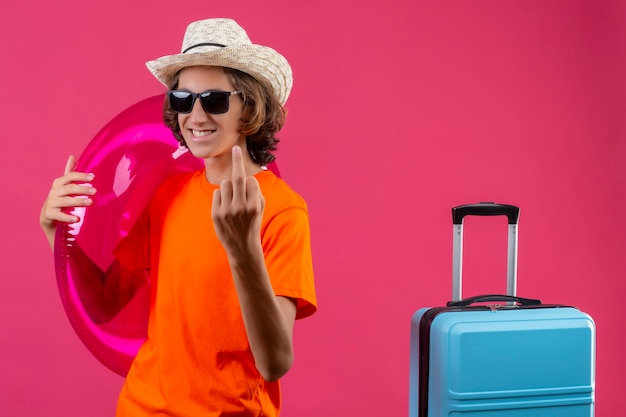 Jonge knappe kerel in oranje t-shirt en zomerhoed die zwarte zonnebril draagt die opblaasbare ring houdt die middelvinger positief en gelukkig status met reiskoffer toont over roze achtergrond