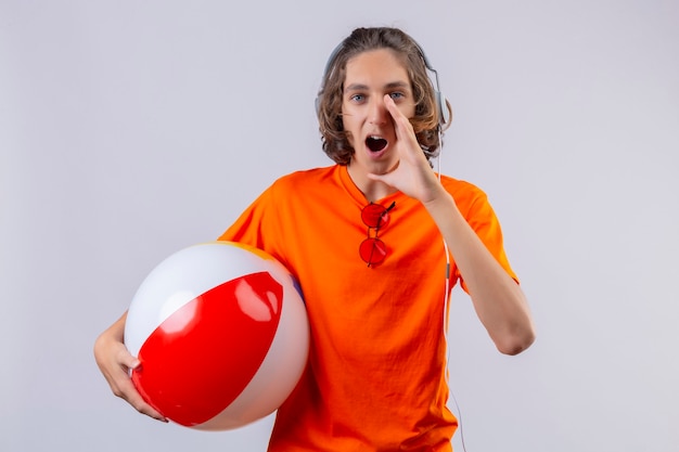 Jonge knappe kerel in oranje t-shirt met koptelefoon houden opblaasbare bal schreeuwen of iemand bellen met hand in de buurt van mond op zoek verrast staande op witte achtergrond
