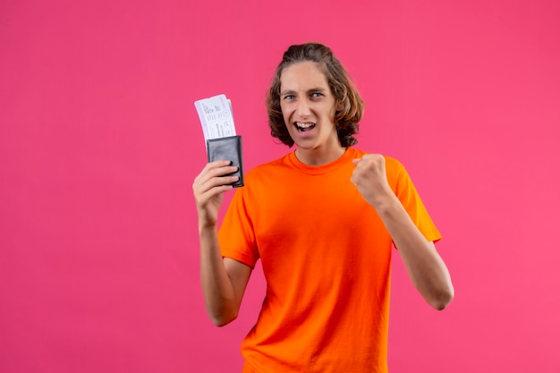 Jonge knappe kerel in oranje t-shirt met luchtkaartjes die vuist het glimlachen vrolijk winnaarconcept verhogen die zich over roze achtergrond bevinden