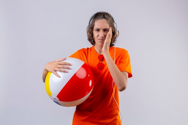 Jonge knappe kerel in oranje t-shirt met opblaasbare bal met koptelefoon op zoek onwel aanraken van wang met kiespijn staande op witte achtergrond