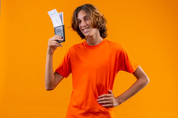 Jonge knappe kerel in oranje t-shirt met vliegtickets die camera kijken met vertrouwen glimlach staande