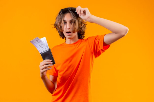 Jonge knappe kerel in oranje t-shirt met vliegtickets die zijn bril uitstellen en kijkt verbaasd en verbaasd over gele achtergrond