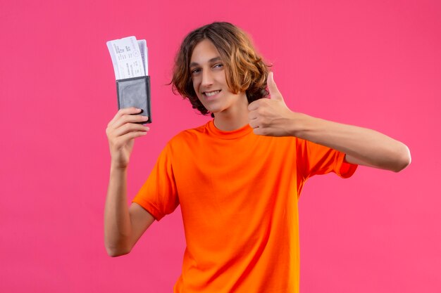 Jonge knappe kerel in oranje t-shirt met vliegtickets op zoek zelfverzekerd duimen opdagen glimlachend vrolijk staande over roze achtergrond