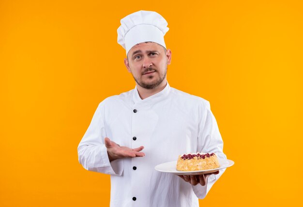 Jonge knappe kok in chef-kok uniform bedrijf en wijzend op plaat van cake geïsoleerd op oranje ruimte