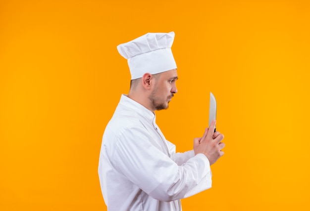 Jonge knappe kok in chef-kok uniform bedrijf mes kijken naar het staande in profiel bekijken op geïsoleerde oranje ruimte