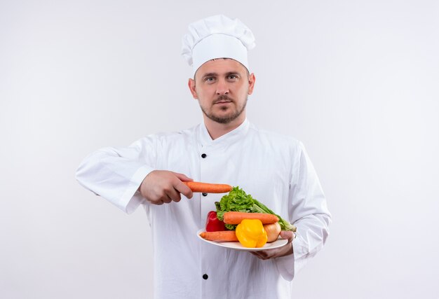 Jonge knappe kok in chef-kok uniforme plaat met groenten te houden en naar hen met wortel te richten die op geïsoleerde witte ruimte kijken