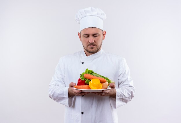 Jonge knappe kok in chef-kok uniforme plaat met groenten te houden en ze op geïsoleerde witte ruimte te bekijken