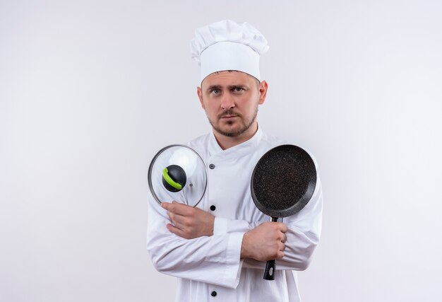 Jonge knappe kok in de koekenpan van de chef-kok eenvormige holding en pannendeksel op zoek geïsoleerd op witte ruimte