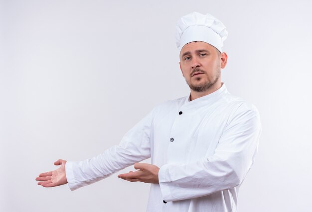 Jonge knappe kok in eenvormige chef-kok die met handen aan kant richt die geïsoleerd op witte ruimte kijken