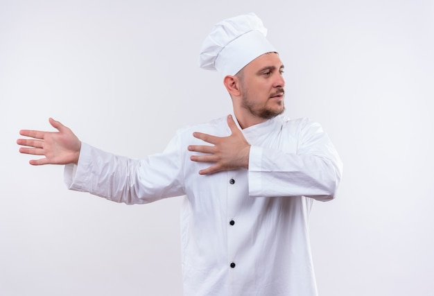 Jonge knappe kok in eenvormige chef-kok die met handen aan linkerkant richt en kant op geïsoleerde witte ruimte bekijkt