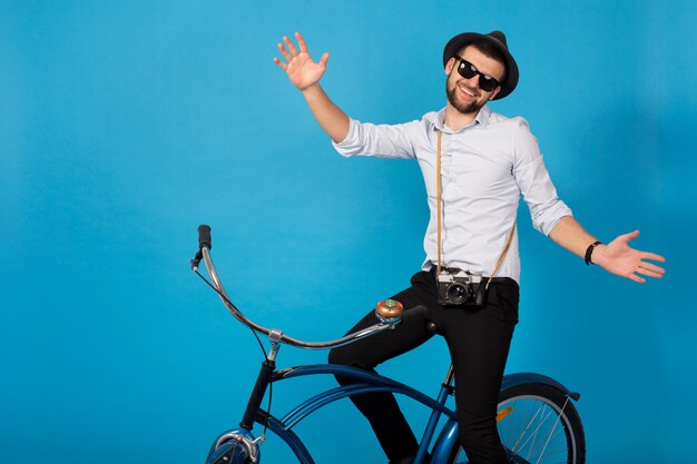 Jonge knappe lachende gelukkig man reizen op hipster fiets, poseren op blauwe studio achtergrond, shirt, hoed en zonnebril dragen