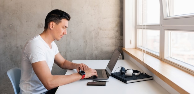 Jonge knappe lachende man in casual outfit zittend aan tafel bezig met laptop alleen thuis blijven