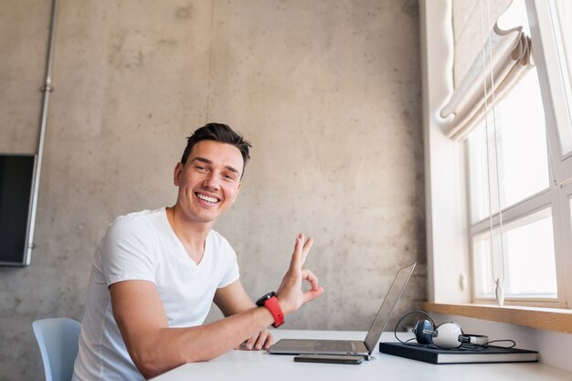 Jonge knappe lachende man in casual outfit zittend aan tafel bezig met laptop, freelancer thuis