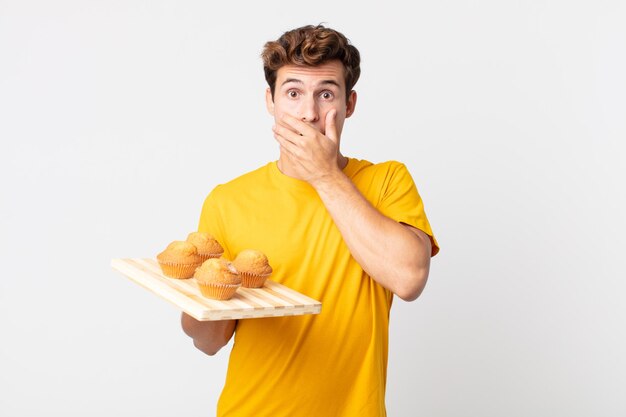 Jonge knappe man die mond bedekt met handen met een geschokte man met een dienblad met muffins Premium Foto