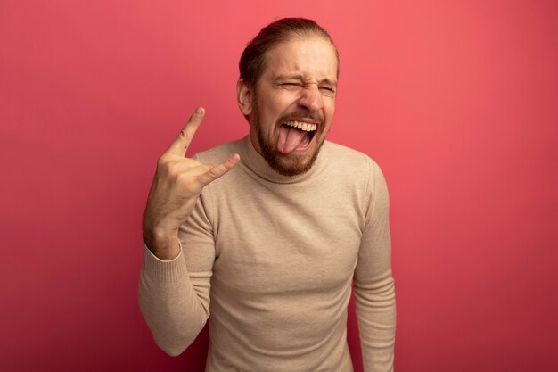 Jonge knappe man in beige coltrui gek gelukkig tong uitsteken waardoor rock symbool staande over roze muur