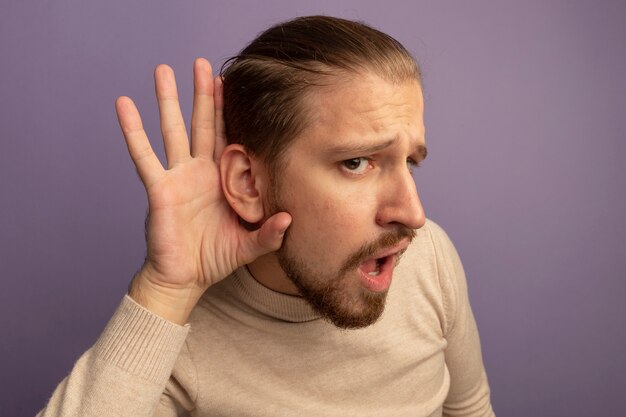 Jonge knappe man in beige coltrui holdng hand dichtbij zijn oor die roddels probeert te luisteren die zich over lila muur bevinden