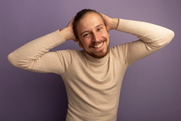 Jonge knappe man in beige coltrui kijken naar voorkant lachend met blij gezicht met handen op zijn hoofd staande over lila muur