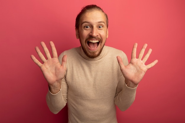 Gratis foto jonge knappe man in beige coltrui kijken voorkant blij en opgewonden met open handpalmen staande over roze muur