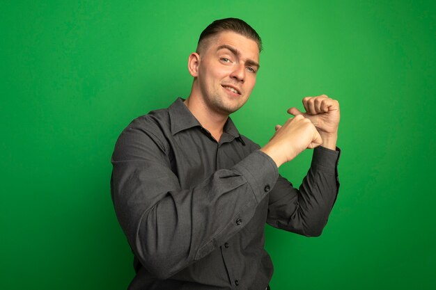 Jonge knappe man in grijs overhemd glimlachend zelfverzekerd wijzend met vingers terug staande over groene muur