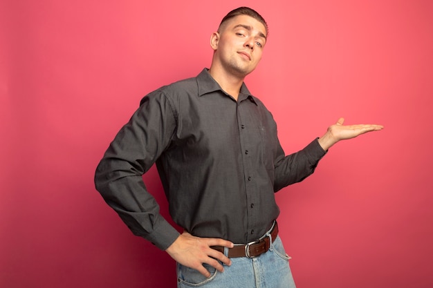 Jonge knappe man in grijs shirt iets presenteren met arm van zijn hand op zoek zelfverzekerd staande over roze muur