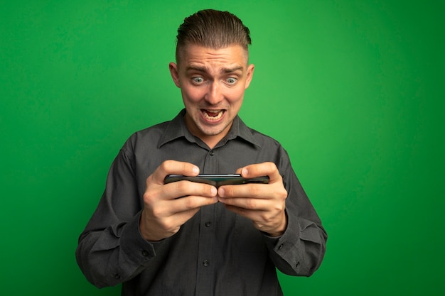 Jonge knappe man in grijs shirt met behulp van smartphone spelen opgewonden en emotionele staande over groene muur