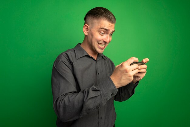 Jonge knappe man in grijs shirt met smartphone blij en opgewonden staande over groene muur