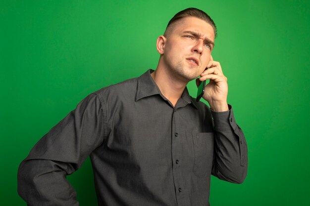 Jonge knappe man in grijs shirt praten op mobiele telefoon met ernstig gezicht staande over groene muur
