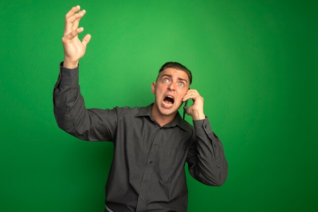 Jonge knappe man in grijs shirt schreeuwen met boze uitdrukking met opgeheven hand tijdens het praten op mobiele telefoon staande over groene muur