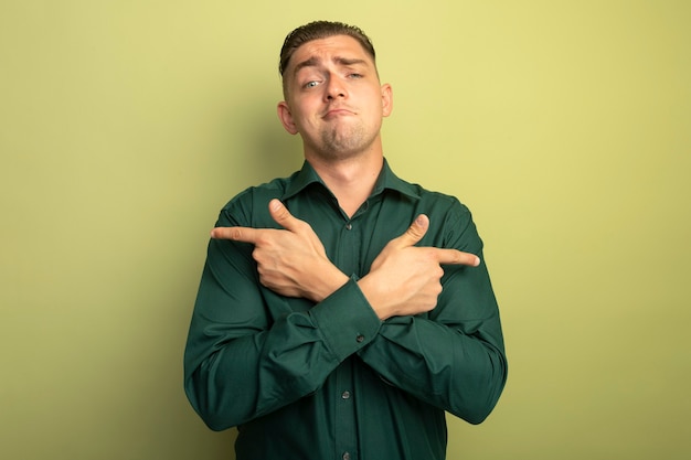 Jonge knappe man in groen overhemd met zelfverzekerde uitdrukking die de handen kruist die met de wijsvingers naar de andere kanten wijzen