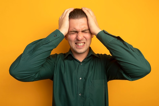 Gratis foto jonge knappe man in groen shirt met hand op zijn hoofd lijdt aan sterke hoofdpijn staande over oranje muur