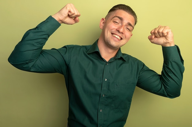 Jonge knappe man in groen shirt vuisten gelukkig en positief winnaar concept staande boven lichte muur opheffen