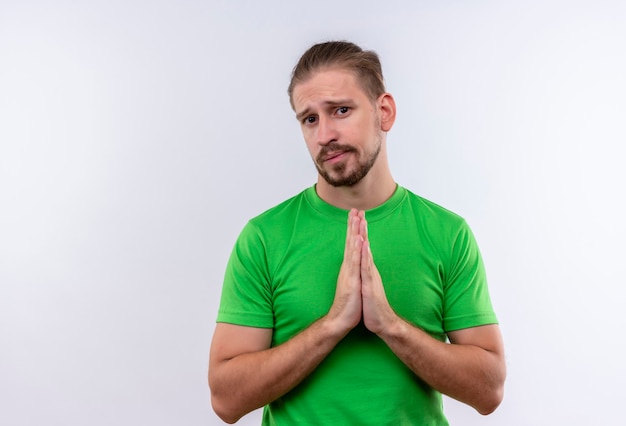 Jonge knappe man in groen t-shirt bedelen en bidden met handen samen emotioneel en bezorgd staande op witte achtergrond