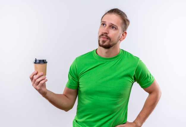 Jonge knappe man in groen t-shirt met kopje koffie opzij kijken ontevreden staande op witte achtergrond