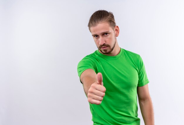 Jonge knappe man in groen t-shirt op zoek zelfverzekerd tonen aan camera duimen omhoog staan op witte achtergrond