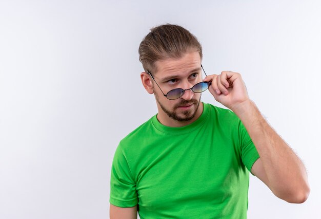 Jonge knappe man in groen t-shirt opzij kijken zijn bril met zelfverzekerde uitdrukking op gezicht staande over witte achtergrond opstijgen