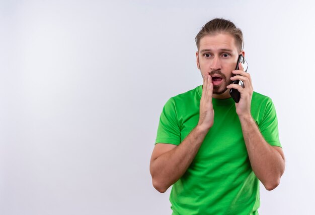 Jonge knappe man in groen t-shirt praten over mobiele telefoon op zoek verrast en verbaasd staande op witte achtergrond