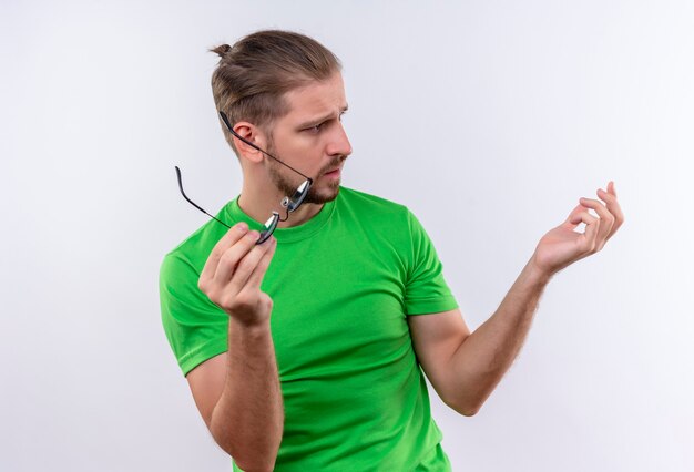 Jonge knappe man in groene t-shirt met zonnebril opzij kijken met verwarren uitdrukking op gezicht staande op witte achtergrond