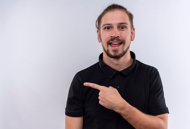 Jonge knappe man in zwart poloshirt glimlachend vrolijk wijzend naar de kant staande op witte achtergrond