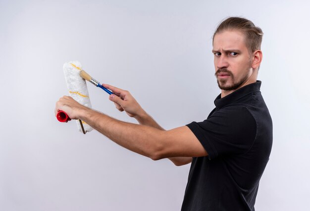 Jonge knappe man in zwart poloshirt met verfroller en kwast opzij kijken fronsend staande op witte achtergrond