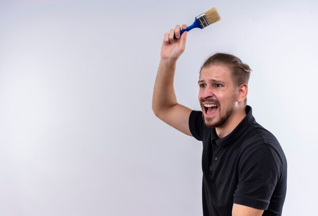 Jonge knappe man in zwart poloshirt zwaaien met een kwast met agressieve uitdrukking staande op witte achtergrond