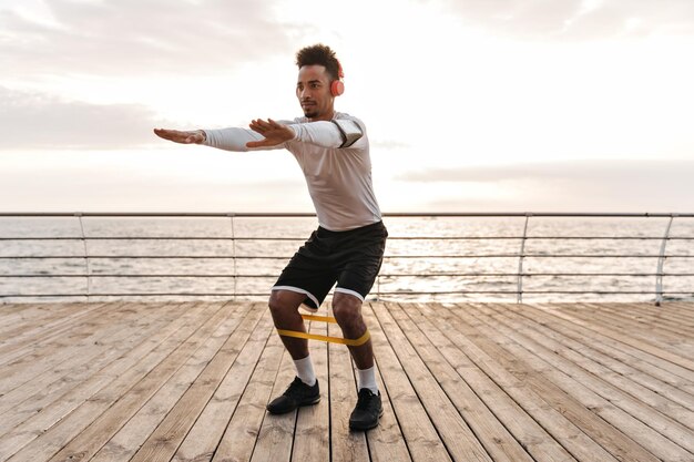 Jonge knappe man in zwarte korte broek en wit t-shirt luistert naar muziek in koptelefoons en squats met fitnessrubber in de buurt van zee