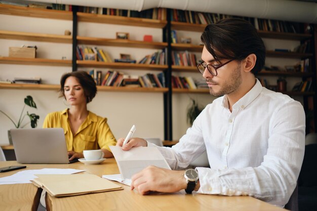 Jonge knappe man met een bril die aan tafel zit en zorgvuldig werk plant met zijn collega op de achtergrond in een modern kantoor