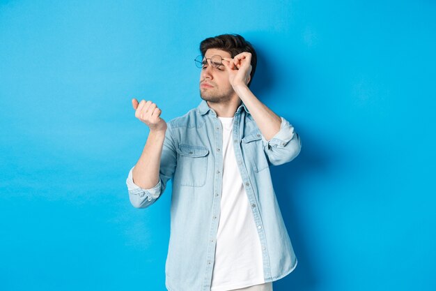 Jonge knappe man met een bril die naar zijn vingernagels kijkt, manicure controleert, over blauwe achtergrond staat